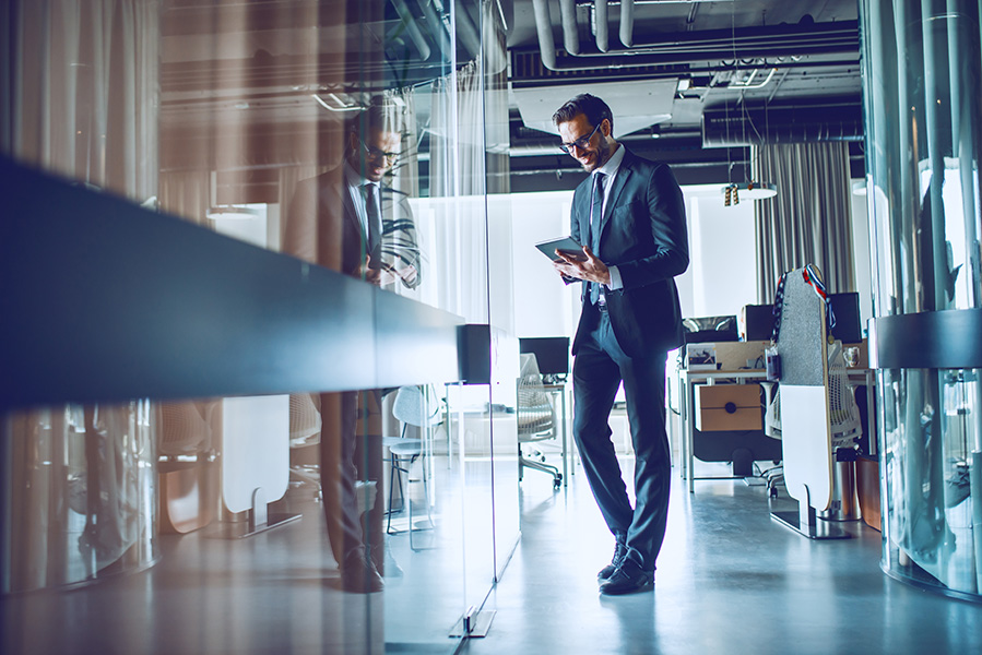 Full length of attractive elegant smiling Caucasian businessman in suit and with eyeglasses standing in front of his office and using tablet.